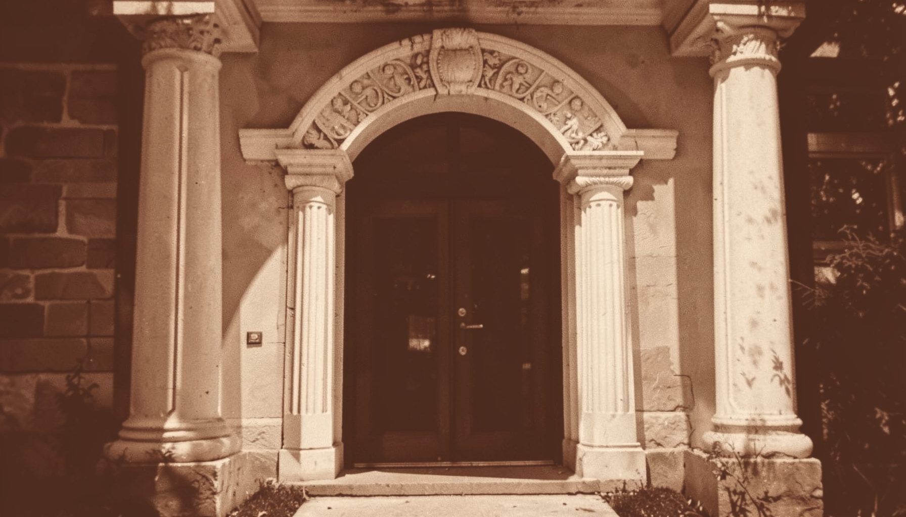 A warm duotone photograph of a softly lit lodge doorway with classical columns and a carved stone arch, rendered in deep brown and cream tones, conveying tradition and quiet invitation.